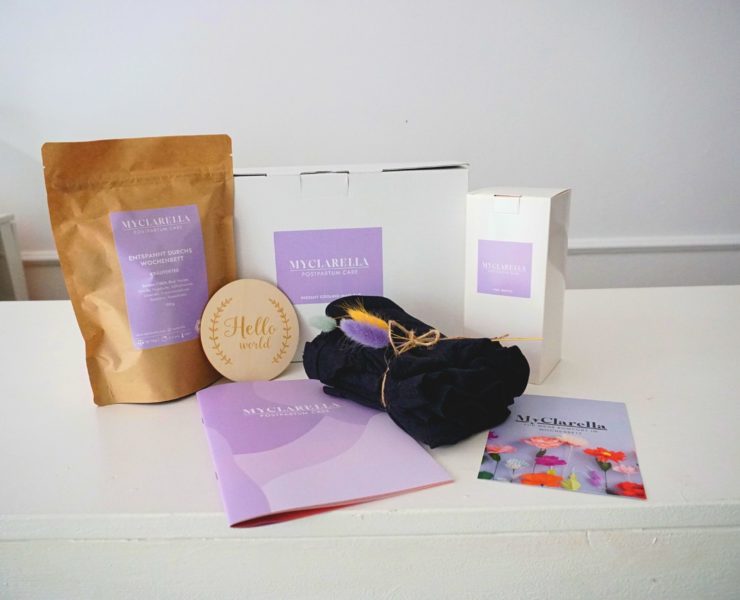 Die drei grundlegenden Wochenbett-Helferlein: Kühlbinden, elastische Unterwäsche und “Peri Bottle" mit Wochenbett-Kräutertee und einen dekorative Holzscheibe mit der Aufschrift “Hello World”.